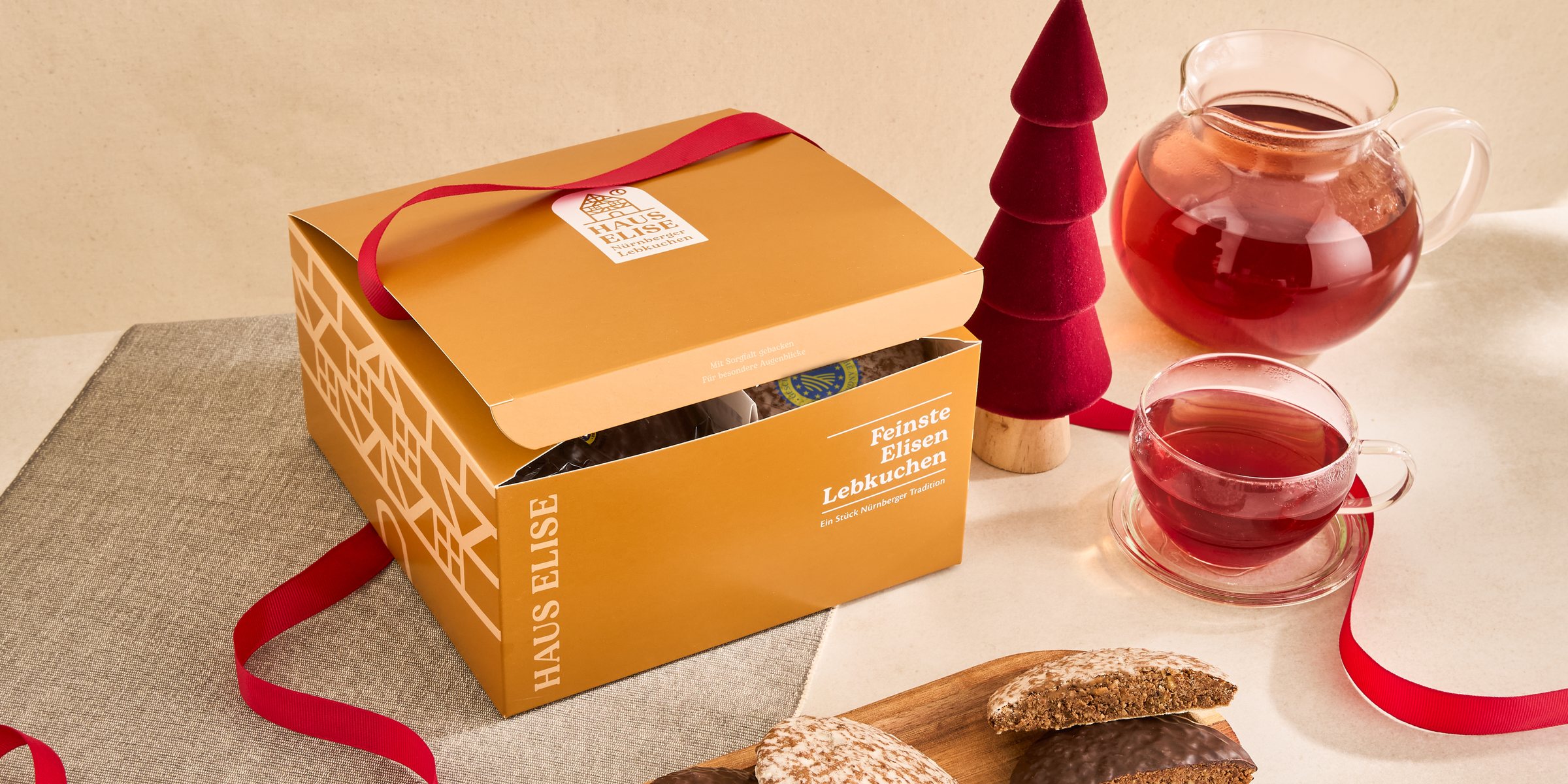 HAUS ELISE Geschenkbox mit rotem Band, daneben eine Kanne und eine Tasse Tee sowie schokolierte und glasierte Elisen-Lebkuchen auf einem Holzbrett